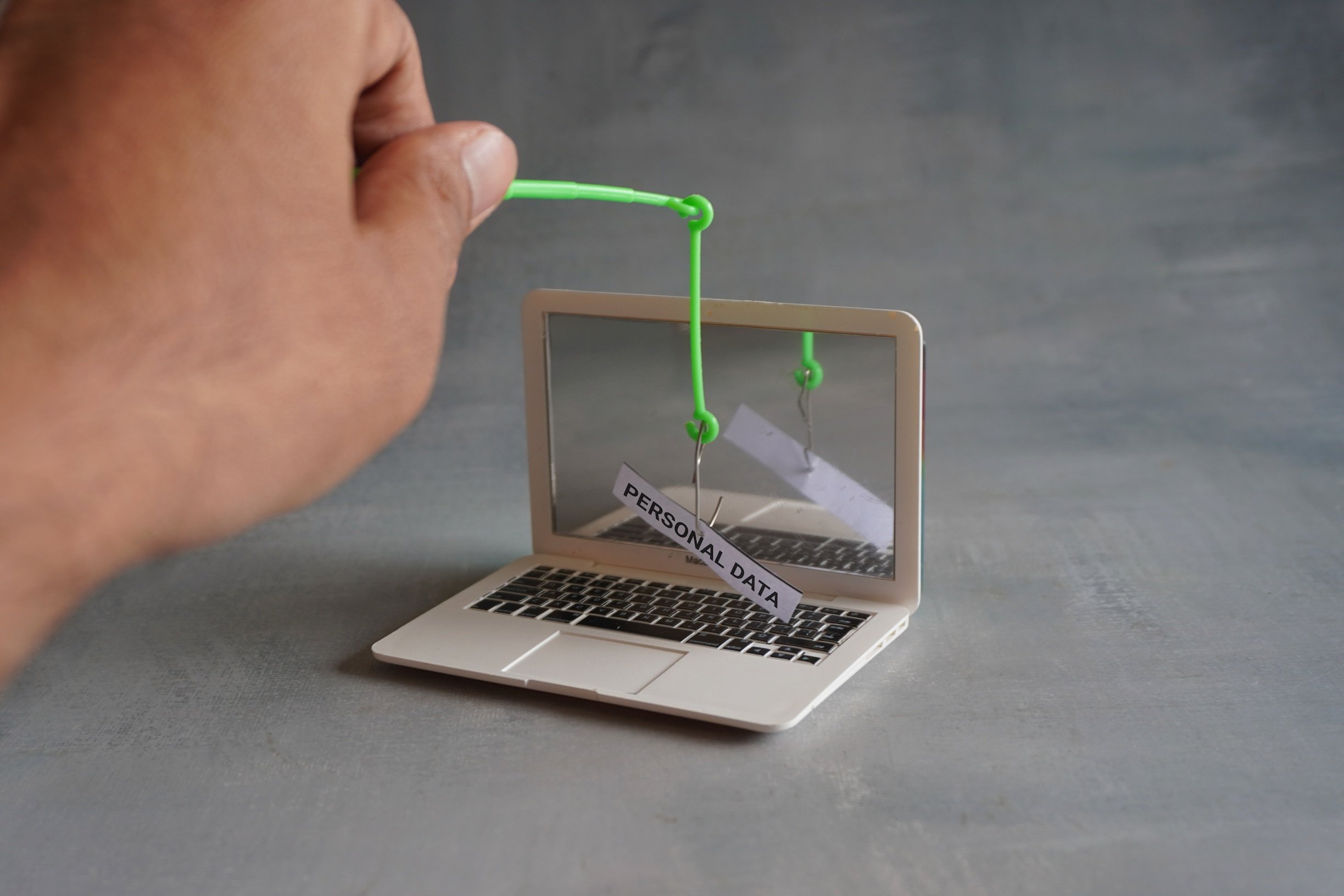 A hand uses a fishing hook and line to pull a "personal data" note through a toy laptop screen, perfectly symbolizing the dangers of phishing—not fishing. A hand uses a fishing hook and line to pull a "personal data" note through a toy laptop screen, perfectly symbolizing the dangers of phishing—not fishing.