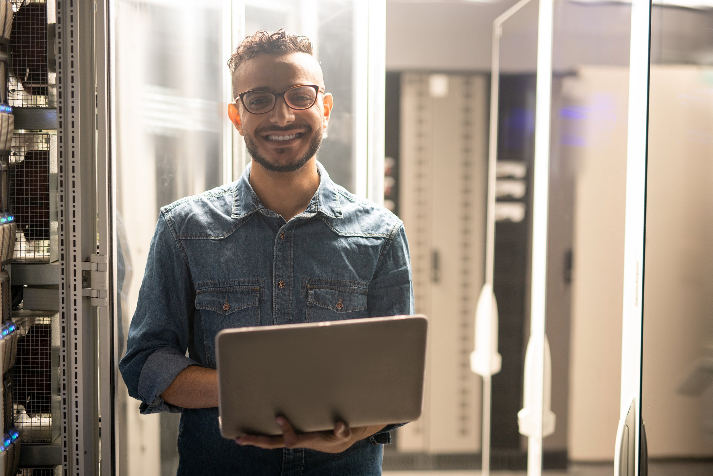 Portrait of cheerful satisfied young middle-eastern server engineer in glasses standing in datacenter room and using laptop