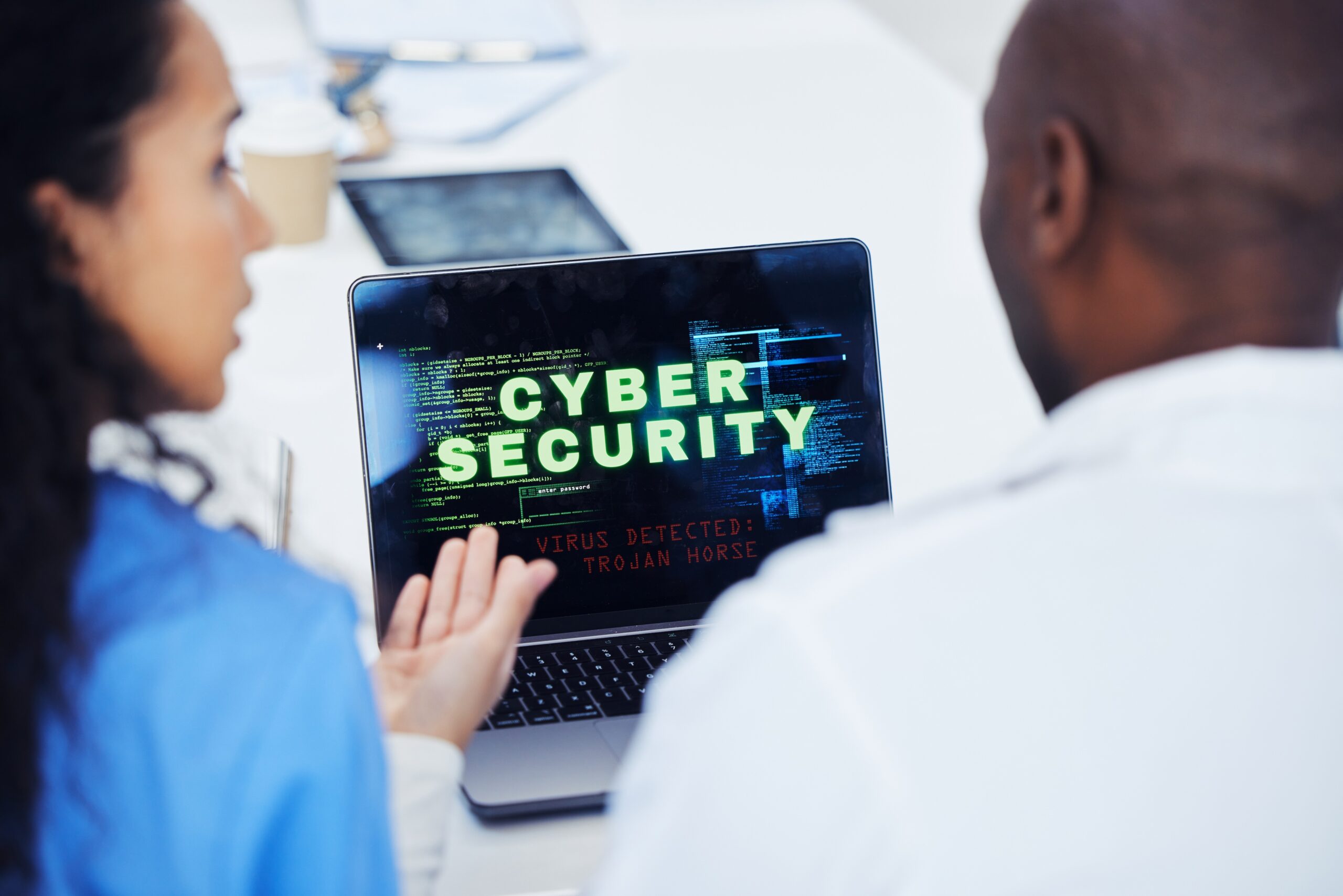 Two people discuss a laptop screen displaying "CYBER SECURITY" and "VIRUS DETECTED: TROJAN HORSE" in a bright, modern office setting. Cybersecurity concept