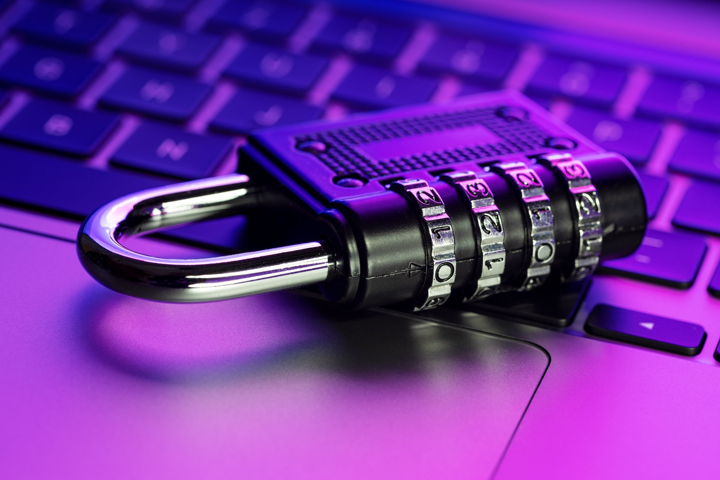 A combination lock rests on a laptop keyboard, illuminated by purple lighting, symbolizing cybersecurity.