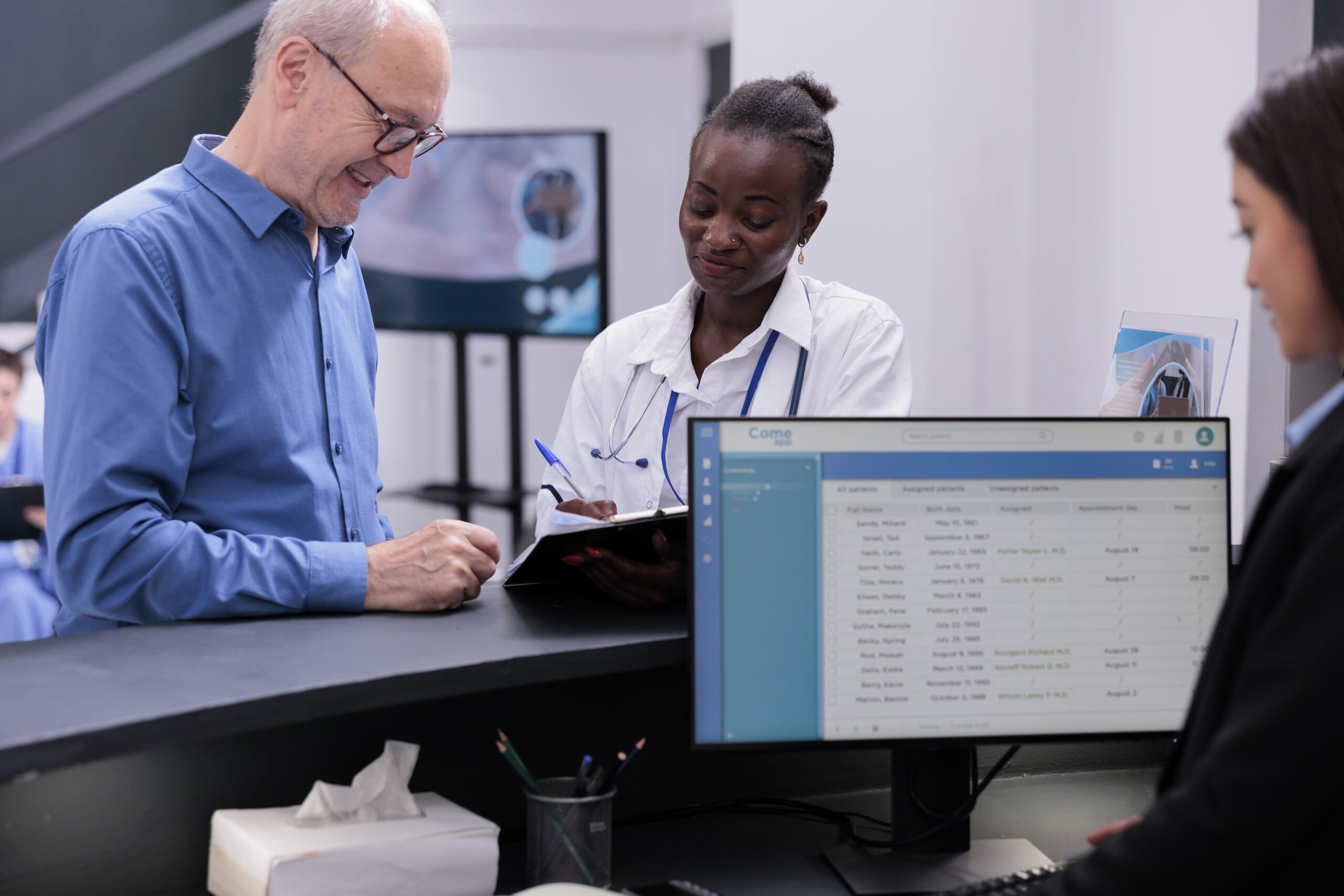 Man in blue shirt and doctor standing at a reception desk, discussing a clipboard. Computer monitor on the desk displays scheduling software. HIPPA compliance concept