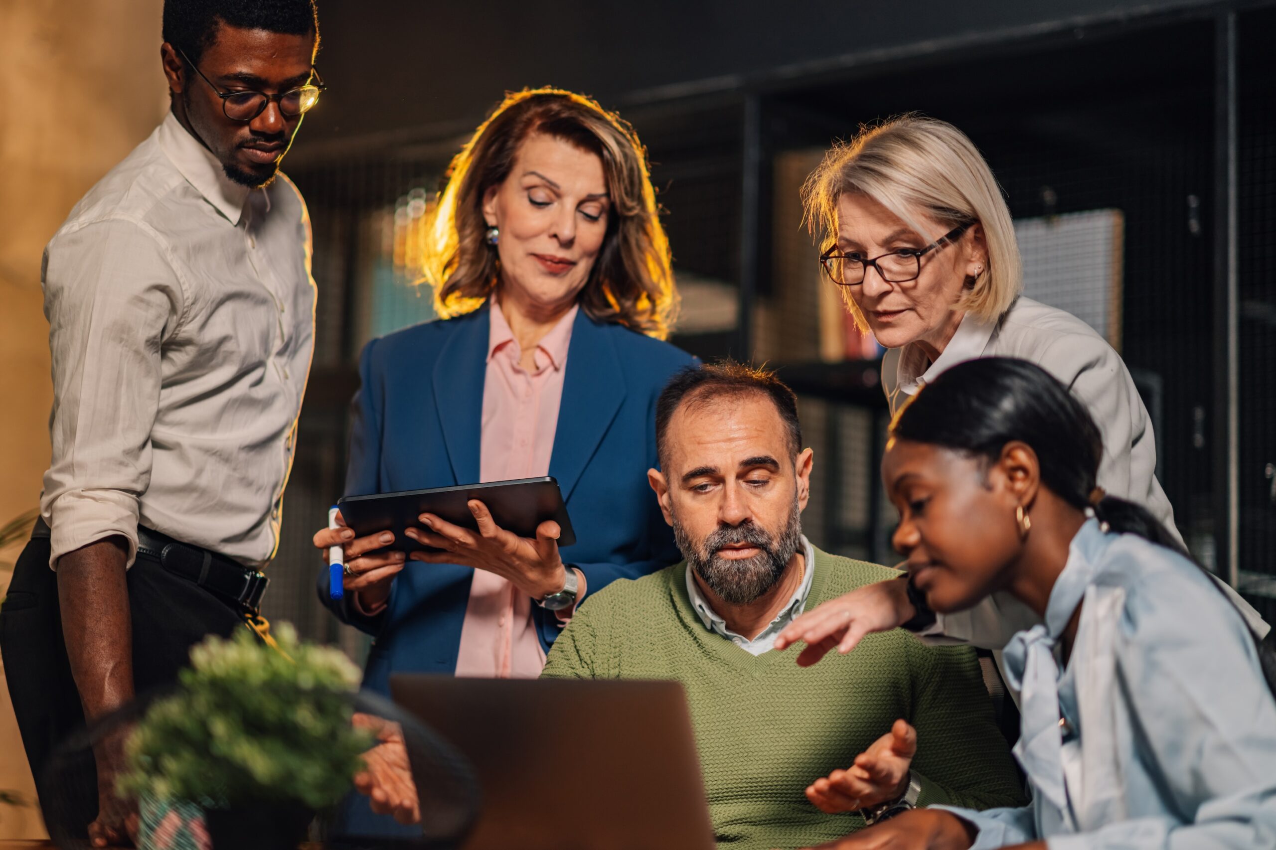 Five people gather around a laptop, engaged in discussion. One person holds a tablet. The setting appears to be an office. Co-Managed IT vs. Fully Managed IT Services Concept