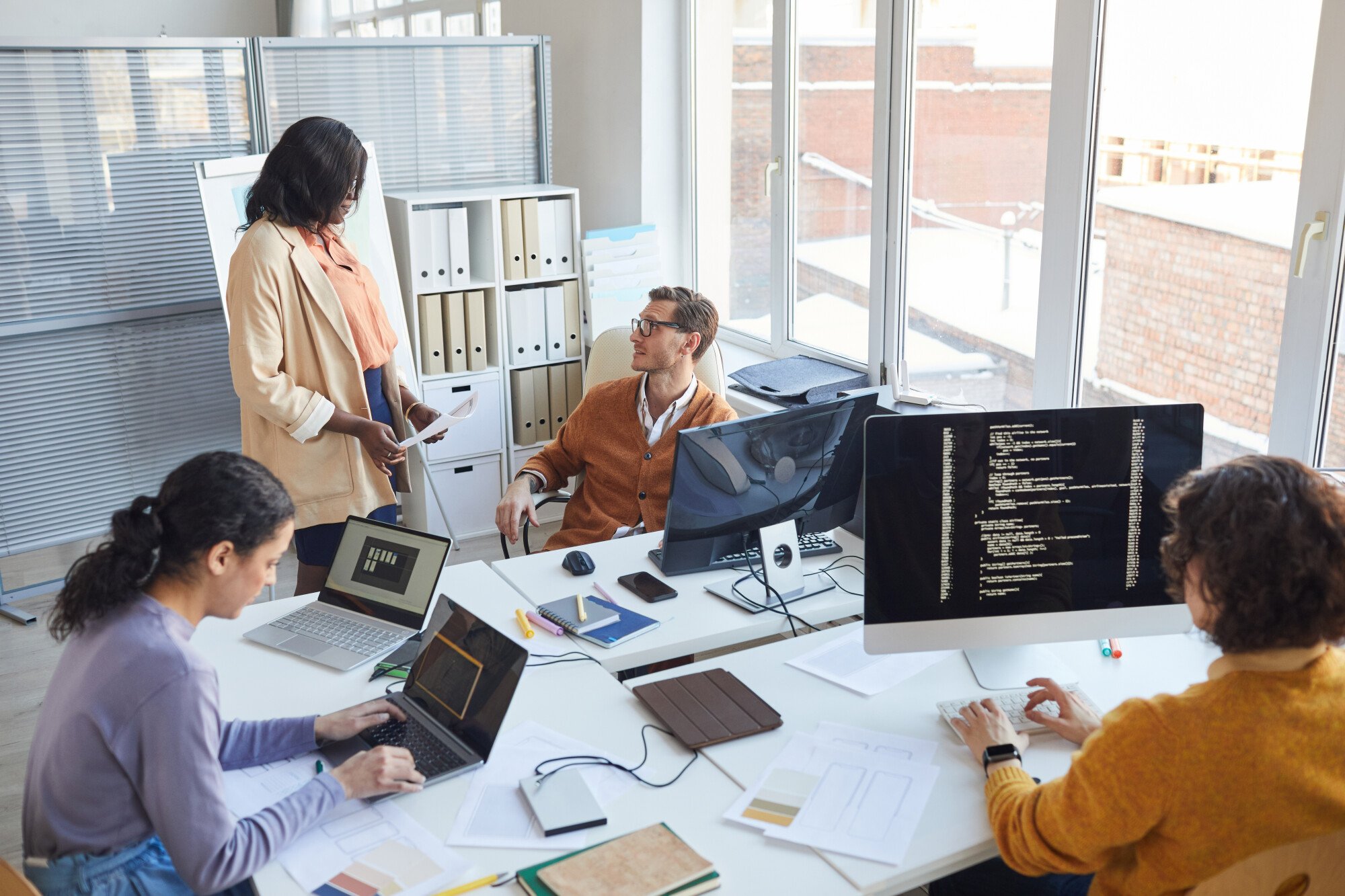 A group of people gather around a desk, focused on a person using a laptop.