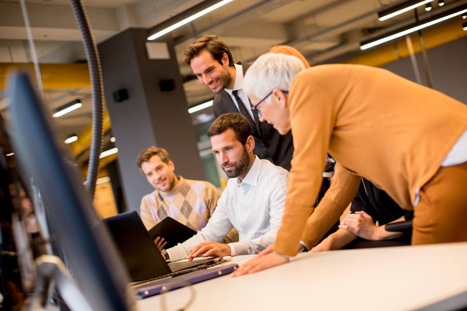 A group of people gather around a desk, focused on a person using a laptop.