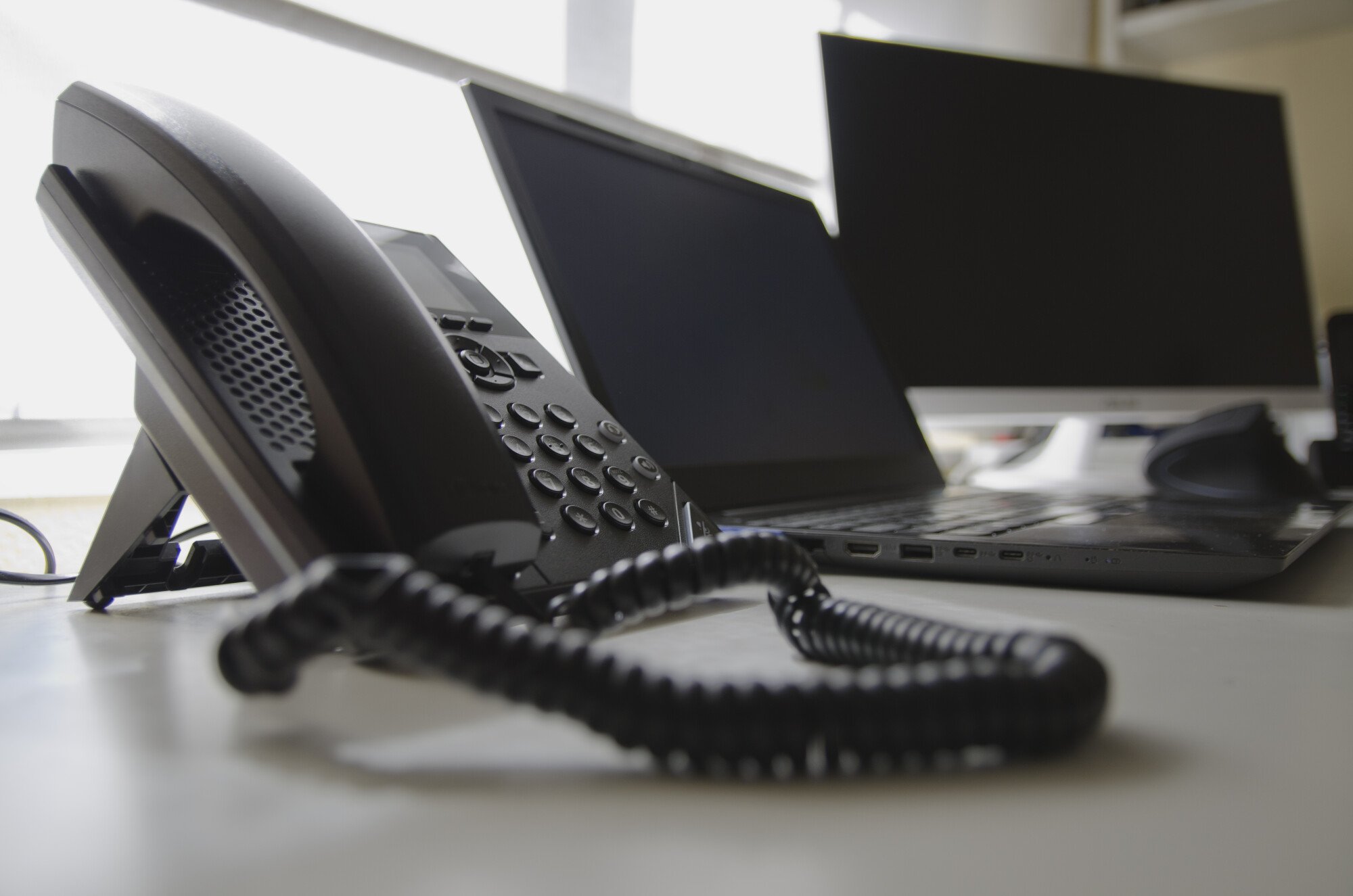 Office desk with a corded phone, two laptops, and a monitor, arranged against a window with natural light.