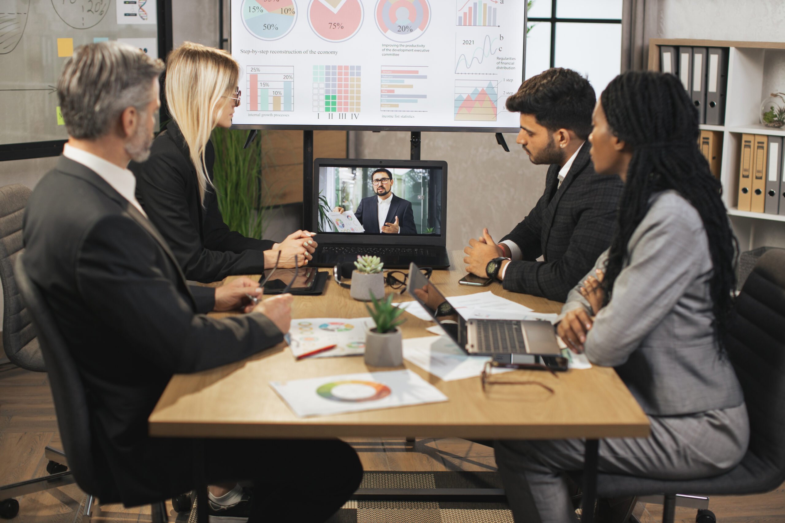 Four people in business attire sit around a table in a meeting room, viewing charts on a screen and a video call with a man. Laptops, papers, and charts are on the table. Four people in business attire sit around a table in a meeting room, viewing charts on a screen and a video call with a man. Laptops, papers, and charts are on the table.