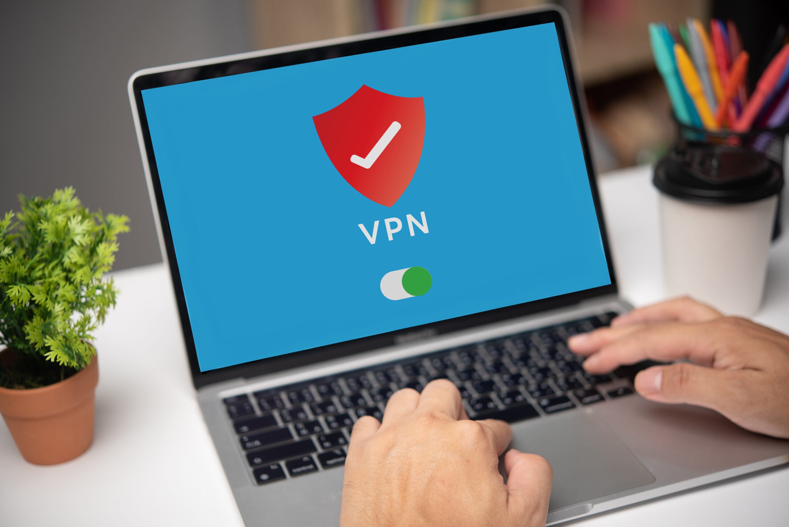 A person types on a laptop displaying a VPN shield icon with a toggle switch, highlighting that traditional VPNs and firewalls are no longer effective. Nearby are a coffee cup, plant, and colorful pens.