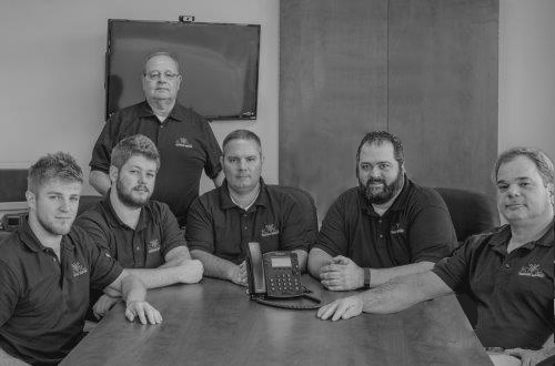 Six men in matching polo shirts sit and stand around a conference table with a speakerphone, posing for a group photo in an office setting.